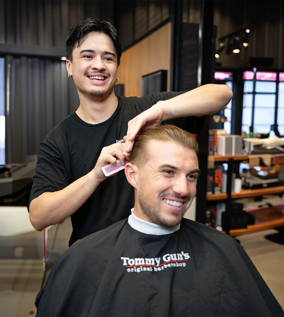 Photo of a man getting a haircut at a Tommy Gun's Original Barbershop, both the barber and man are smiling.