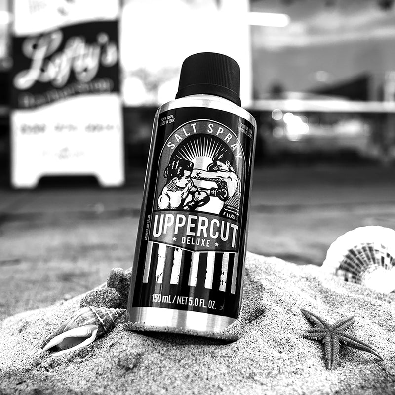 A photo of Uppercut branded Salt Spray, sitting on a staged hill of sand with a beach theme.